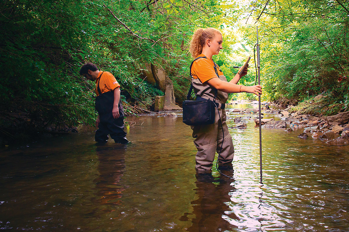 A woman stands in a creek taking a scientific measurement.