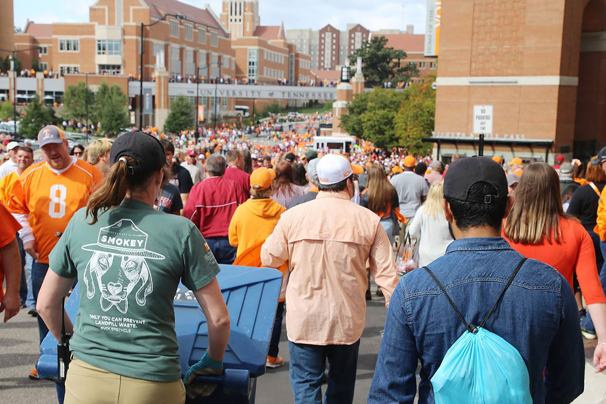 A waste warrior pushes a recycle bin through game day crowd.